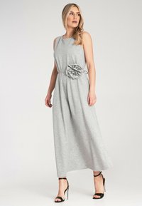 Szara, bezrękawnikowa sukienka maxi z teksturowanym, falbaniastym detalem w formie kwiatu w talii, wykonana z miękkiego materiału, w połączeniu z czarnymi sandałami na wysokim obcasie.