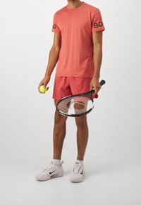 Tenue de sport corail comprenant une chemise à manches courtes et un short, tenant une raquette de tennis et une balle. Baskets blanches avec des accents noirs.