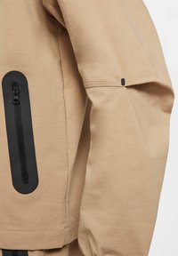 Casaco bege com uma textura suave, apresentando um bolso com fechozip preto e detalhes pretos nas mangas. Design simples com linhas limpas.