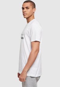 Hvid bomulds t-shirt med korte ærmer, rund hals og trykt tekstdetalje i sort. Modellen står i profil mod en neutral baggrund.