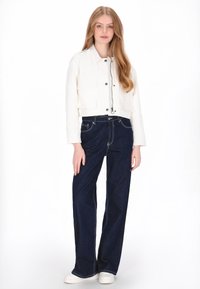Witte cropped jas met voorzakken, donkerblauwe jeans met wijde pijpen en witte sneakers; gladde textuur en contrasterende stikseldetails.