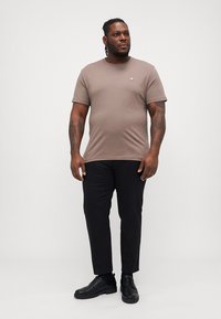 Tricou scurt maro cu mânecă scurtă, guler rotund și un logo mic, asortat cu pantaloni negri și pantofi negri. Are o siluetă ajustată.