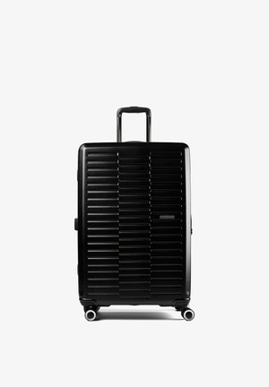 Valigia rigida nera con scanalature orizzontali, manico telescopico esteso, quattro ruote rotanti e logo American Tourister sul davanti.