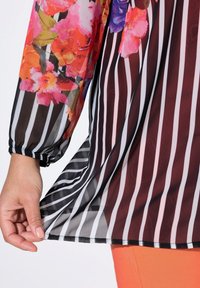 Tunic trasparente floreale con colori vivaci e strisce verticali in bianco e nero, con polsini arricciati e un tessuto leggero e fluido.