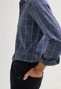 Chemise à carreaux bleu marine avec un col boutonné, présentant des lignes blanches en quadrillage ; manches longues retroussées jusqu'aux coudes, associée à un jean foncé.