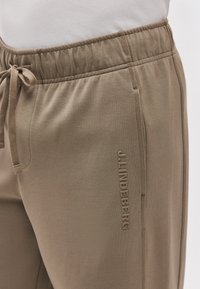 Κοντινό πλάνο μπεζ φόρμας με ελαστική μέση και κορδόνι, με κάθετο λογότυπο JLINDENBERG στην πλευρά.