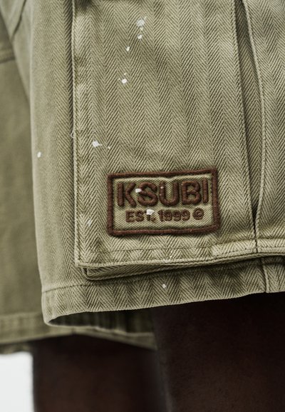 Βερμούδα από ύφασμα καμηλό με μοτίβο ψαροκόκαλο, λευκές πιτσιλιές μπογιάς και καφέ ταμπέλα με την επιγραφή "KSUBI Est. 1999 ©" σε πόδι ατόμου.
