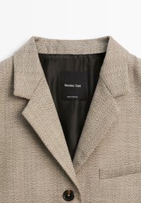 Béžový texturovaný blejzr s klopami, jedním černým knoflíkem a vnitřní etiketou s nápisem "Massimo Dutti", vyrobený v Portugalsku.