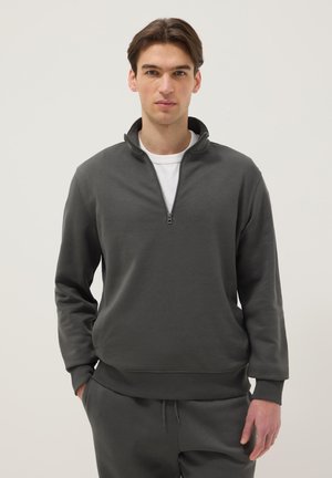 Jeune homme portant un sweat-shirt à demi-fermeture éclair gris foncé sur un t-shirt blanc avec un pantalon gris foncé assorti, debout devant un fond clair uni.
