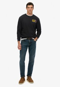 Sudadera negra con cuello redondo y gráfico frontal, combinada con jeans azules y botas marrones. Diseño sencillo, textura y ajuste casual.