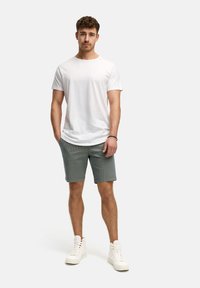 Tricou alb cu mânecă scurtă, asortat cu pantaloni scurți gri texturat. Modelul poartă adidași înalți albi, stând pe un fundal neutru.