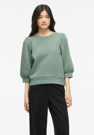 Femme portant un haut vert sauge à manches bouffantes trois-quarts et un pantalon noir droit, debout devant un fond blanc.