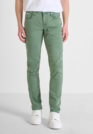 Pantalones de mezcla de algodón verde con un corte ajustado, diseño de cinco bolsillos y cierre de botones. Combinados con zapatillas blancas.