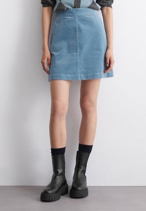 Kobieta w jasnoniebieskiej mikrospódniczce z sztruksu, czarnych skarpetkach i masywnych czarnych botkach, stojąca na tle białej ściany.