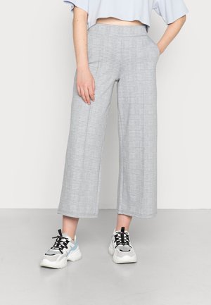 Person iført lysegrå ternede vidde cropped bukser med lommer og hvide chunky sneakers med sorte snørebånd på en ensfarvet baggrund.