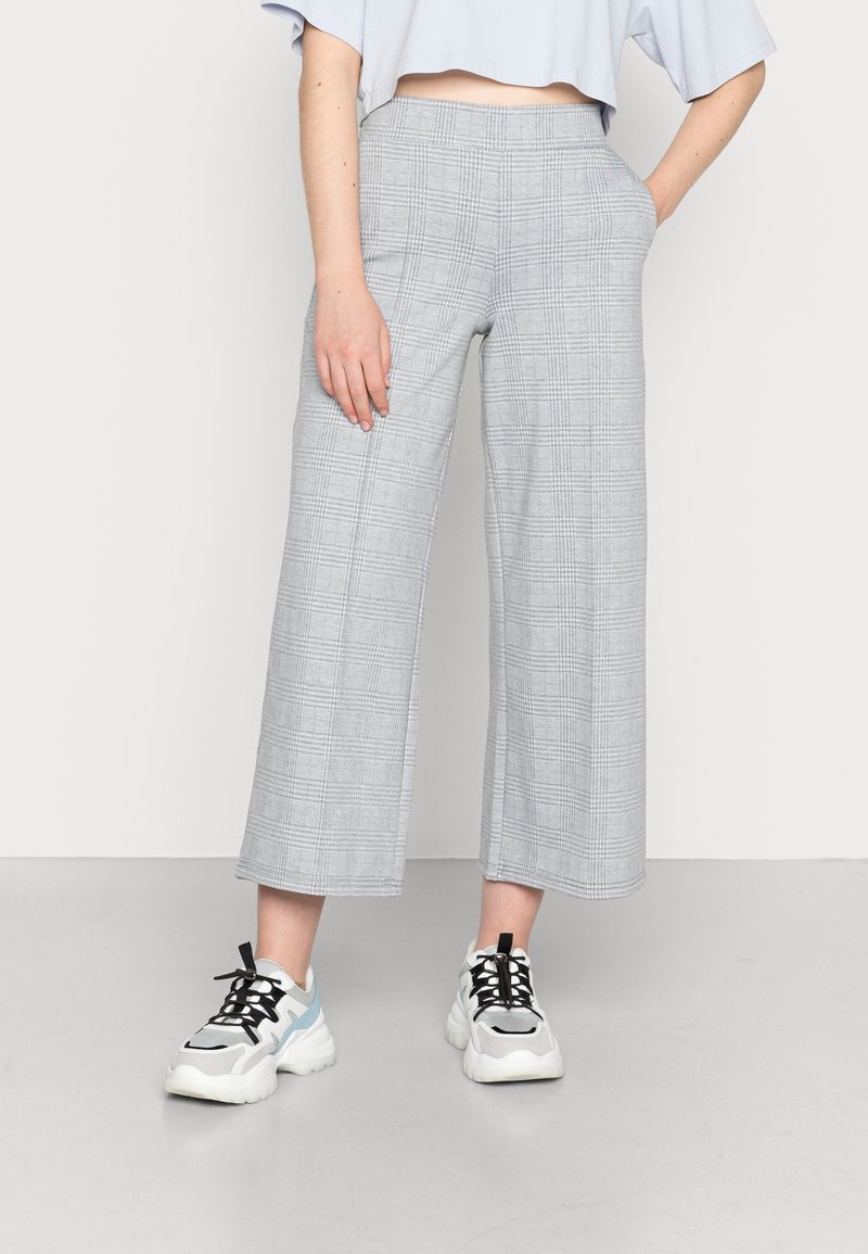 Personne portant un pantalon large court à carreaux gris clair avec poches et des baskets blanches épaisses avec lacets noirs sur un fond uni.