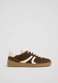 Brown suede sneaker with a cream leather accent and rubber sole. Features a low-top design with white laces and subtle side patterns.