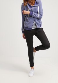 Felpa con cappuccio viola, dotata di zip, indossata sopra una maglietta a righe, abbinata a leggings neri e sneaker bianche. Tessuto morbido, vestibilità casual.