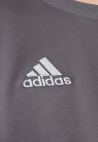 Grå atletisk skjorte lavet af struktureret stof med et hvidt broderet Adidas-logo på brystet, kendetegnet ved en rund halsudskæring.