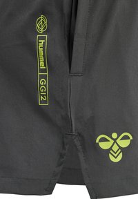 Grå shorts i lättviktstyg har klara gröna logotyper: "hummel"-text och en stiliserad bi-design nära fållen.