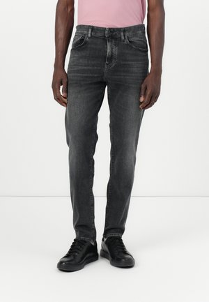 Mand iført falmede sorte slim-fit jeans, sorte sneakers og en lyserød T-shirt stoppet ind i bukserne foran hvid baggrund.