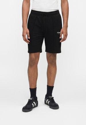 Άνδρας που φοράει μαύρα σορτς με κορδόνι και το λογότυπο "JJ Studio" στο αριστερό μηρό, μαύρες κάλτσες και μαύρα αθλητικά παπούτσια με λευκές ρίγες.