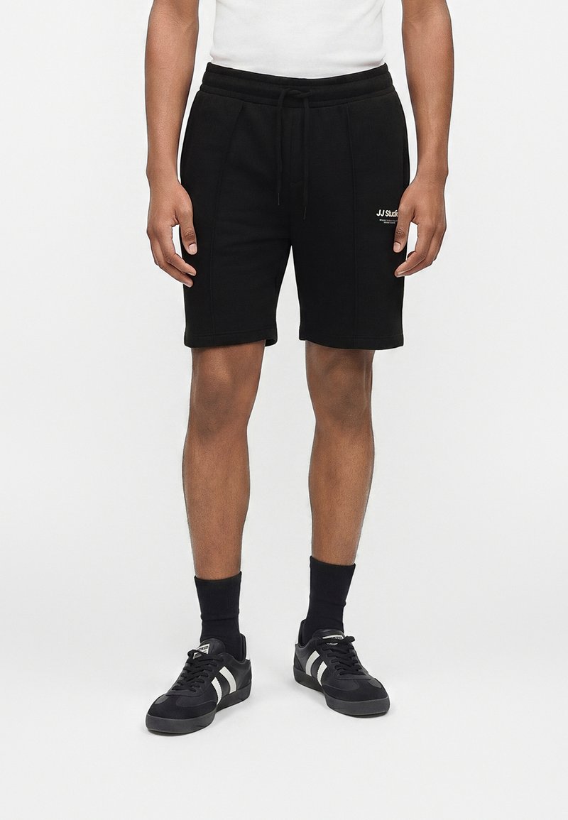 Άνδρας που φοράει μαύρα σορτς με κορδόνι και το λογότυπο "JJ Studio" στο αριστερό μηρό, μαύρες κάλτσες και μαύρα αθλητικά παπούτσια με λευκές ρίγες.