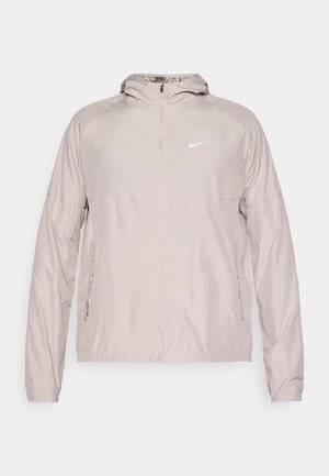 Lys beige Nike-jakke i lett materiale, med frontglidelås, hette og sidelommer. Reflekslogo på brystet.