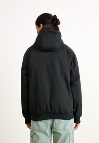 Black hooded jacket with ribbed cuffs and hem. Smooth fabric texture and simple design. Worn over striped green and white pants.