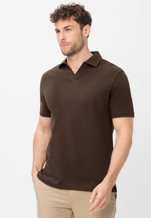 Mann mit lockigem Haar, der ein braunes kurzärmeliges Poloshirt und beige Hose trägt, steht mit einer Hand in der Tasche und schaut leicht nach rechts.