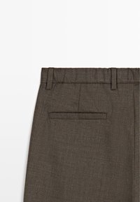 Gros plan sur un pantalon marron sur mesure montrant la taille avec passants pour ceinture et une poche passepoilée arrière unique sur un fond blanc.