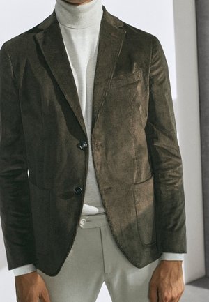 Mężczyzna ubrany w ciemnooliwkową kordurową marynarkę na jasnoszarym golfie i jasnoszarych spodniach, stojący w pomieszczeniu.