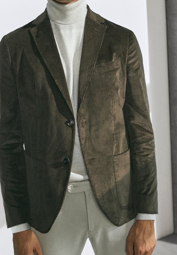 Homme portant un blazer en velours côtelé vert olive foncé sur un pull à col roulé gris clair et un pantalon gris clair, debout à l'intérieur.