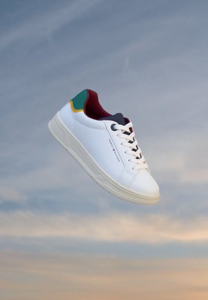 Baskets basses blanches avec des accents verts, jaunes et rouges, des lacets blancs et une semelle beige sur un fond de ciel doux et nuageux.