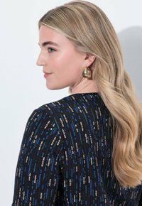Blonde Frau von links mit langen, welligen Haaren, die große goldene Creolen und ein schwarzes Top mit einem blauen und beige gepunkteten Muster trägt.