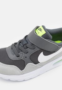 Grå sportskor med mesh- och mockadetaljer, vit Nike-logga, grön accent på tungan och ett kardborreband.