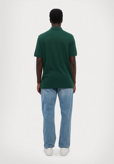 Polo de culoare verde închis cu mâneci scurte, purtat cu blugi albastre deschise și adidași albi, văzut din spate pe un fundal simplu.