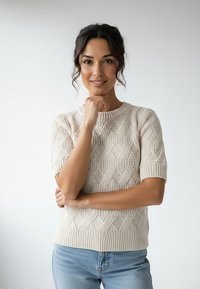 Dzianinowy sweter w kremowym kolorze z teksturowanym wzorem warkocza. Krótkie rękawy i prążkowany dekolt. Noszony z jasnoniebieskimi dżinsami.