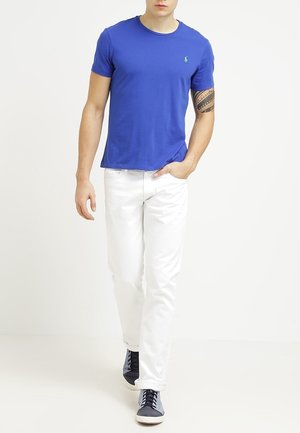 Blue cotton crew-neck t-shirt with a small green logo, paired with white jeans and navy sneakers. Smooth texture, casual fit.
