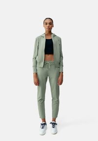 Giacca sportiva zip-up verde chiaro con pantaloni jogger coordinati, top cropped nero sotto e sneakers bianche con lacci blu navy. Tessuto morbido, design aderente.