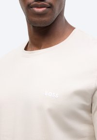 Άνδρας που φοράει ανοιχτό μπεζ μπλουζάκι με λευκό λογότυπο "BOSS" στο στήθος, κοντινή λήψη που δείχνει το κάτω μέρος του προσώπου και τον λαιμό.