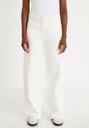 Person iført højtaljede, brede, hvide bukser og hvide sneakers, stående mod en ensfarvet hvid baggrund.