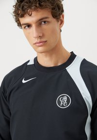Sort sweatshirt med rund hals, hvid Nike-logo og lyseblå akcenter på skuldrene, med et cirkulært CFC-logo vist markant.