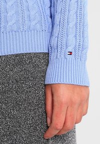 Lichtblauwe kabelgebreide trui met geribde mouwboord, met een klein rood, wit en blauw logo. Gecombineerd met een textuurrijke zwarte kledingstuk.