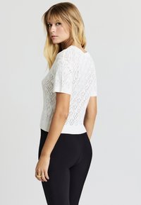 Blonde Frau, die ein weißes, kurzärmliges Strickoberteil mit Diamantmustern und schwarze, enganliegende Hosen trägt und über ihre Schulter schaut.