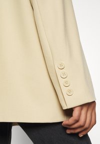 Manche de blazer beige avec quatre boutons assortis, montrée sur le bras d'une personne reposant à côté de son pantalon foncé.