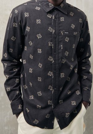 Sort langærmet skjorte med knapper og beige abstrakte mønstre, en forlommelabel med "Volcom", båret med lyse bukser.