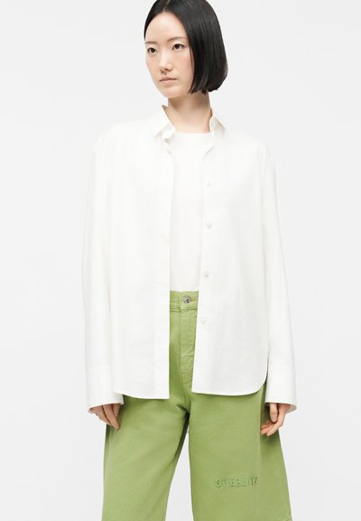 Lühikeste mustade juustega naine, kellel on seljas üleval valge lahtine nööpidega särk, valge topp ja rohelised püksid, millel tikitud "STIEGLITZ".