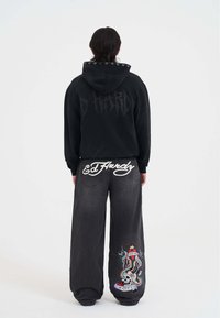 Sudadera negra con capucha, ojales plateados y estampado "D HARD", combinada con jeans grises holgados que presentan el logo blanco de "Ed Hardy" y un colorido diseño de tatuaje.