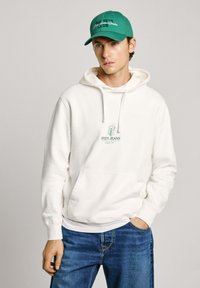 Sudadera blanca con un bolsillo tipo canguro y un logo impreso en verde. Combinada con vaqueros azules. Gorra verde con texto bordado. Atuendo casual.